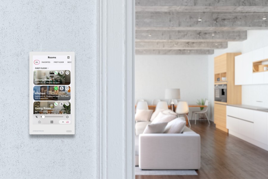 A sleek, wall-mounted smart home control panel displaying room settings, including lighting and temperature controls for the dining room, kitchen, and living room, in a modern, open-concept home with a white kitchen and cozy seating area in the background.