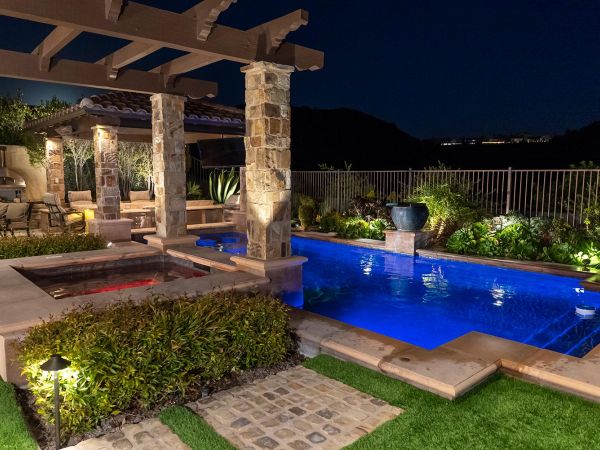 A luxurious outdoor living space at night, featuring a well-lit pool with vibrant blue water, a spa, and a pergola with stone pillars, showcasing the ambiance of a high-end backyard retreat, enhanced by Sonance outdoor audio solutions.