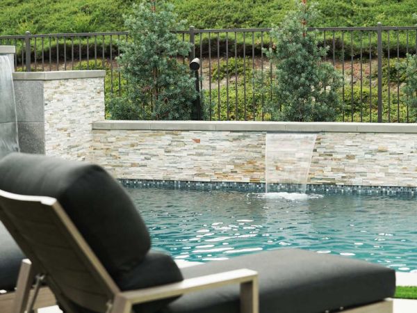 Close-up of a backyard pool with a waterfall feature, framed by a stone wall and evergreen trees, highlighting a serene outdoor space.