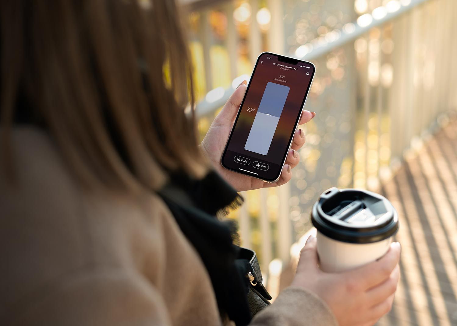 A person holding a smartphone with the Josh AI app open, showing thermostat controls, while holding a coffee cup, with a blurred outdoor background.settings against a plain white background.